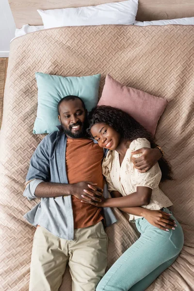 Blick von oben auf afrikanisch-amerikanische Eheleute in lässiger Kleidung, die sich im Bett ausruhen und in die Kamera schauen — Stockfoto