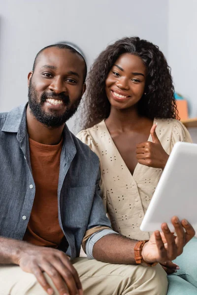 African american man holding digital tablet near wife showing thumb up and looking at camera — Stock Photo