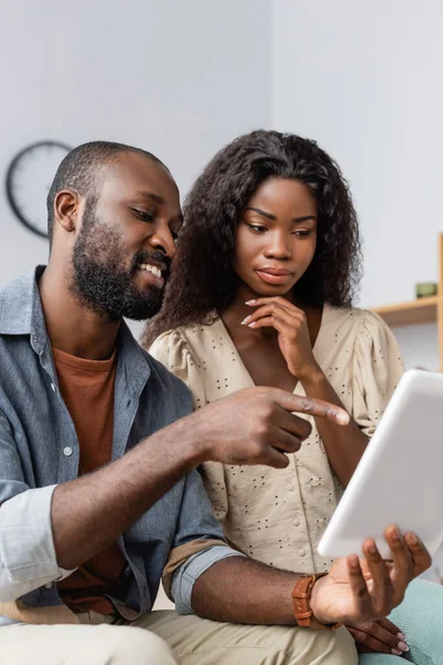 Joven afroamericano hombre apuntando con el dedo a la tableta digital cerca de esposa pensativa - foto de stock