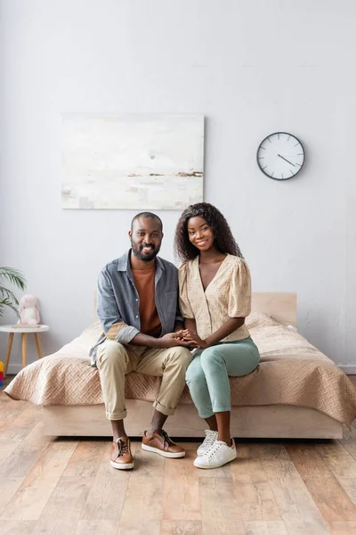 Africano americano marido y esposa en casual ropa cogido de la mano y mirando cámara en dormitorio - foto de stock