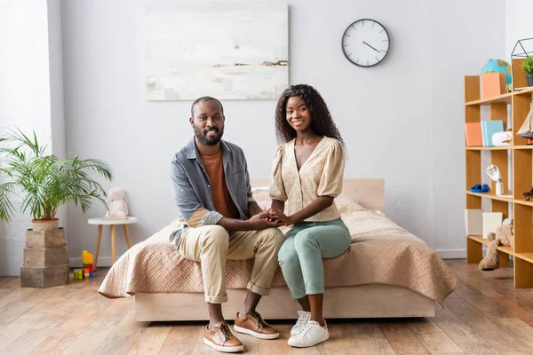 Junges afrikanisch-amerikanisches Paar in lässiger Kleidung, Händchen haltend und auf dem Bett sitzend in die Kamera blickend — Stockfoto