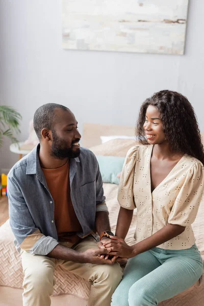 Молода афро-американська пара в повсякденному одязі тримаючись за руки і дивлячись один на одного в спальні. — стокове фото