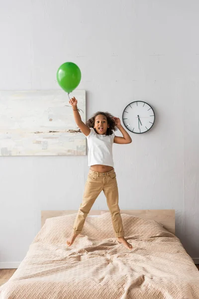Animado afro-americana menina em roupas casuais pulando na cama com balão vermelho — Fotografia de Stock