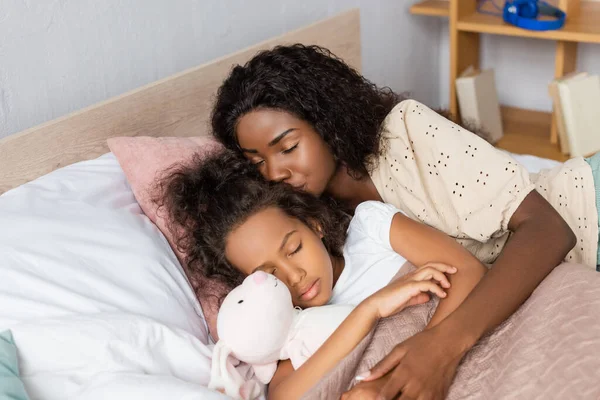 Young african american mother kissing daughter sleeping with toy bunny — Stock Photo