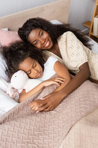 Joven afroamericana americana mujer mirando a cámara mientras acostado cerca durmiendo hija y juguete conejito - foto de stock