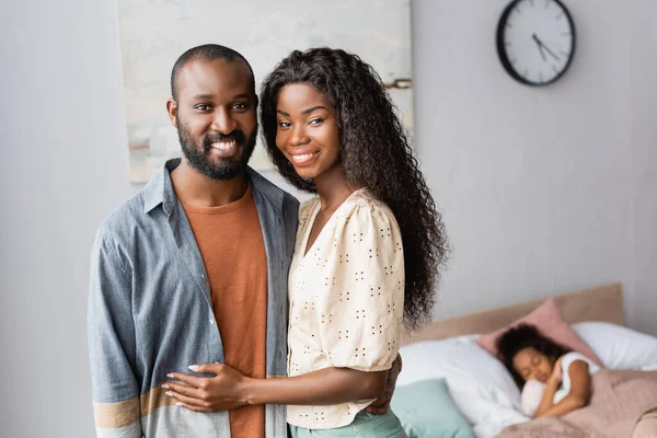 Joven africano americano pareja mirando cámara mientras de pie cerca hija durmiendo en cama - foto de stock