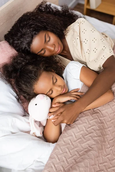 Hochwinkelaufnahme einer afrikanisch-amerikanischen Mutter, die ihre Tochter umarmt, während sie zusammen in der Nähe von Spielzeughasen schläft — Stockfoto