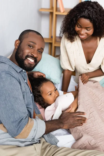 Молодые африканские американские родители сидят рядом с ребенком, лежащим под одеялом с игрушечным кроликом — стоковое фото