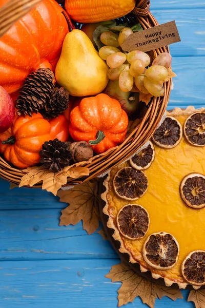 Top view of autumnal harvest in wicker basket with happy thanksgiving card near pumpkin pie on blue wooden background — Stock Photo