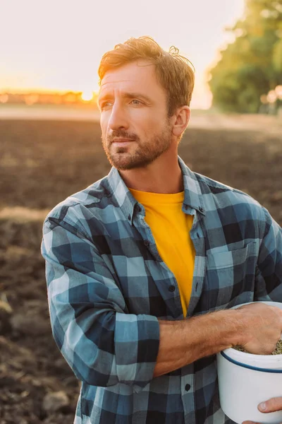 Rancher im karierten Hemd schaut weg, während er mit Eimer auf dem Feld steht — Stockfoto