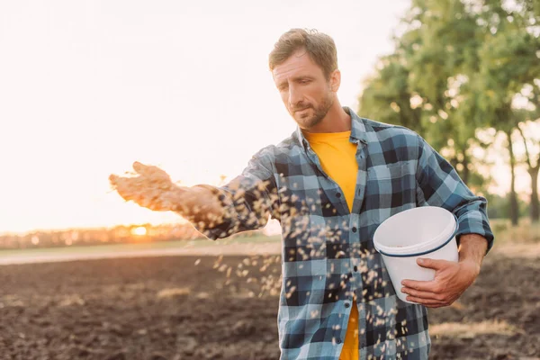 Селективное внимание фермера в клетчатой рубашке, сеющего семена на вспаханном поле — стоковое фото