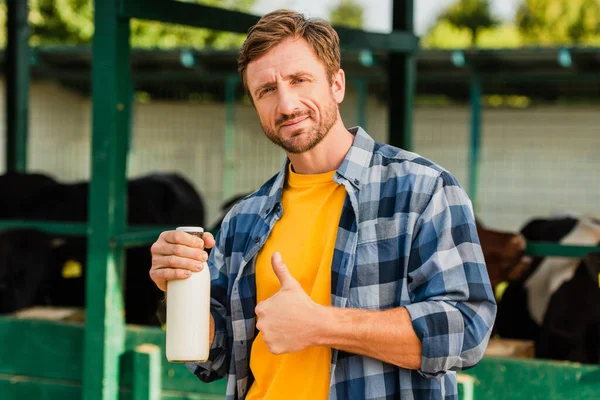 Bauer im karierten Hemd zeigt Daumen hoch, während er eine Flasche frische Milch auf dem Hof hält — Stockfoto