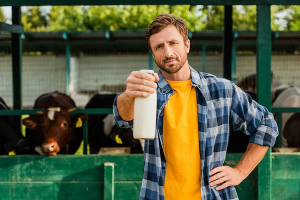 Ranchero en camisa a cuadros de pie cerca de establo con la mano en la cadera y mostrando botella de leche fresca - foto de stock