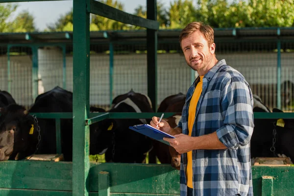 Bauer im karierten Hemd blickt in die Kamera, während er in der Nähe des Kuhstalls steht und auf Klemmbrett schreibt — Stockfoto