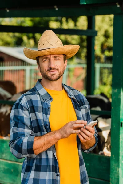 Ранчер в клетчатой рубашке и соломенной шляпе с помощью мобильного телефона и глядя на камеру на ферме — стоковое фото