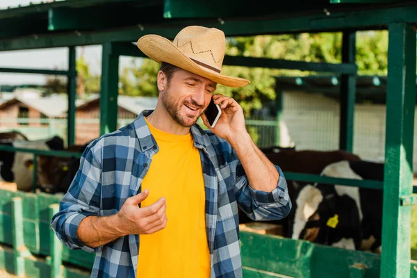 Agricultor en sombrero de paja y camisa a cuadros hablando en el teléfono inteligente cerca de establo en la granja - foto de stock