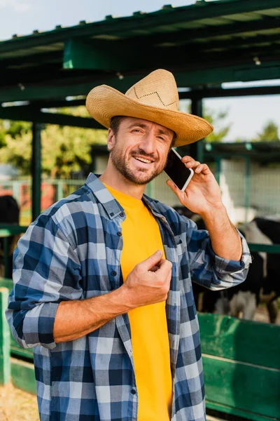 Фермер в соломенной шляпе и клетчатой рубашке разговаривает на смартфоне возле коровника — стоковое фото