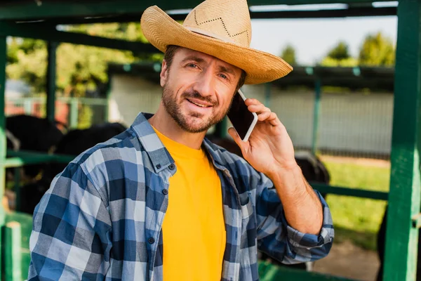 Agricultor en camisa a cuadros y sombrero de paja hablando en el teléfono inteligente mientras mira a la cámara - foto de stock