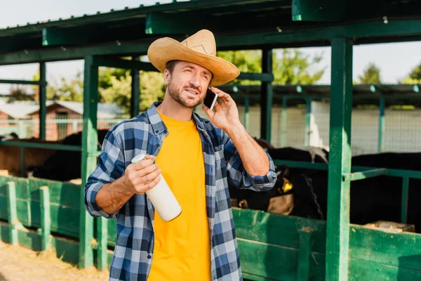 Farmer in checkered shirt and straw hat talking on smartphone while holding bottle of milk — Stock Photo