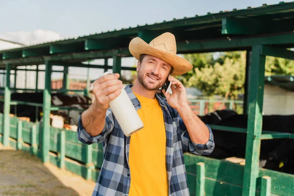 Rancher in straw hat and plaid shirt talking on mobile phone while showing bottle of fresh milk near cowshed — Stock Photo