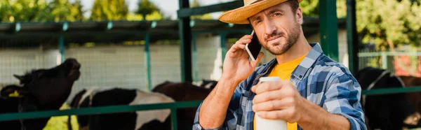 Panoramic concept of farmer in straw hat and plaid shirt holding bottle of milk while talking on mobile phone — Stock Photo