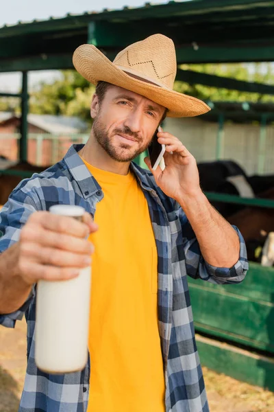 Enfoque selectivo de ranchero en sombrero de paja y camisa a cuadros hablando en el teléfono inteligente mientras que muestra botella de leche - foto de stock