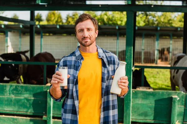 Ranchero en camisa a cuadros mirando a la cámara mientras sostiene la botella y el vaso de leche fresca en la granja - foto de stock