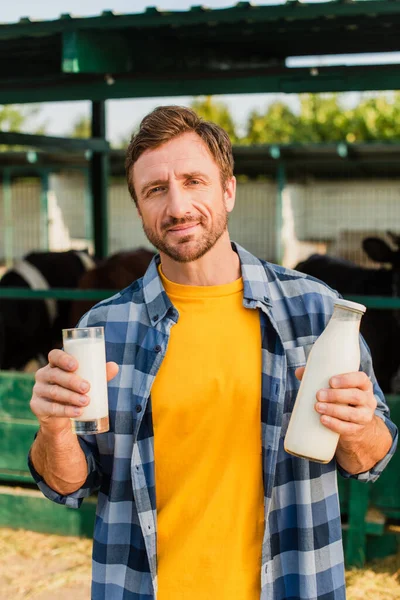 Ranchero en camisa a cuadros sosteniendo botella y vaso de leche fresca en la granja lechera - foto de stock
