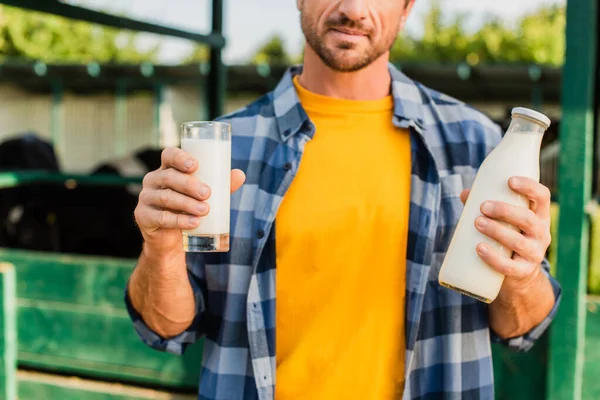 Vista recortada del agricultor en camisa a cuadros sosteniendo botella y vaso de leche fresca cerca de establo - foto de stock