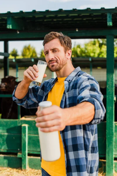 Селективный фокус фермера в клетчатой рубашке, держащего стекло и бутылку молока при взгляде на камеру — стоковое фото