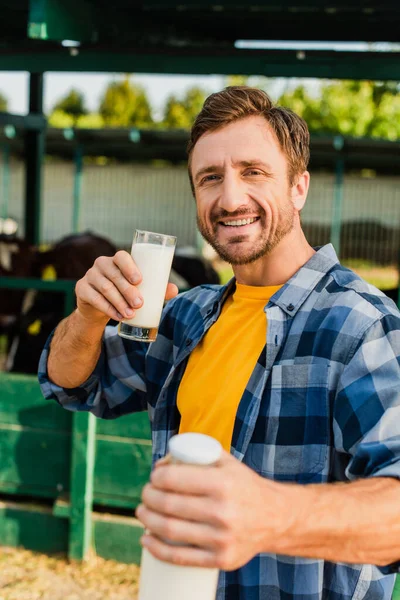 Foco seletivo do fazendeiro na camisa xadrez segurando garrafa e vidro de leite fresco enquanto olha para a câmera — Fotografia de Stock