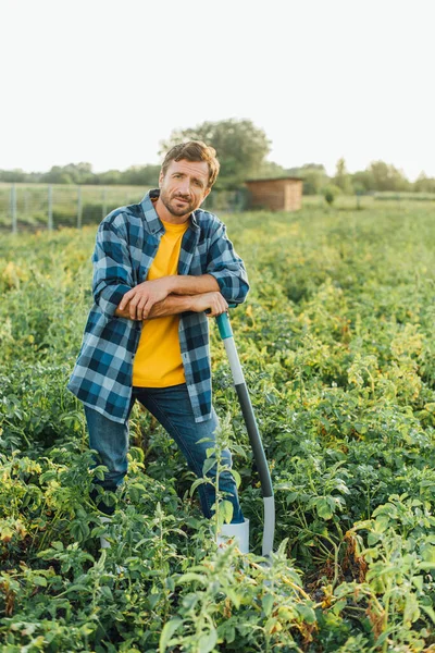 Agriculteur en chemise à carreaux regardant la caméra tout en s'appuyant sur la pelle dans le champ — Photo de stock