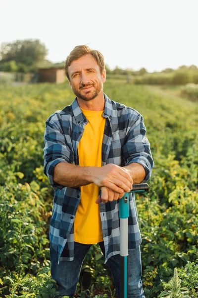 Agriculteur en chemise à carreaux regardant la caméra tout en se tenant debout avec la pelle dans le champ — Photo de stock
