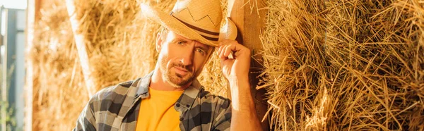 Farmer near bale of hay touching straw hat while looking at camera, website header — Stock Photo