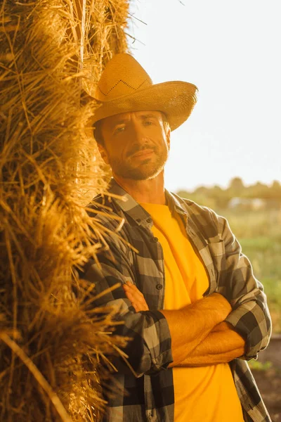 Rancher en chemise à carreaux et chapeau de paille regardant la caméra tout en s'appuyant sur une balle de foin avec les bras croisés — Photo de stock