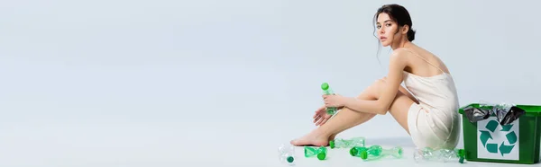 Concept panoramique de femme brune en robe de soie assise près des bouteilles en plastique et poubelle avec panneau de recyclage sur blanc — Photo de stock