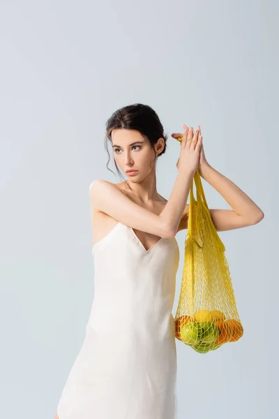 Morena mujer en vestido de seda sosteniendo bolsa de hilo con frutas maduras en blanco, concepto de ecología - foto de stock