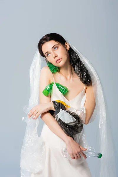 Femme avec sac en plastique sur la tête debout en robe de soie avec des bouteilles vides isolées sur gris, concept écologie — Photo de stock