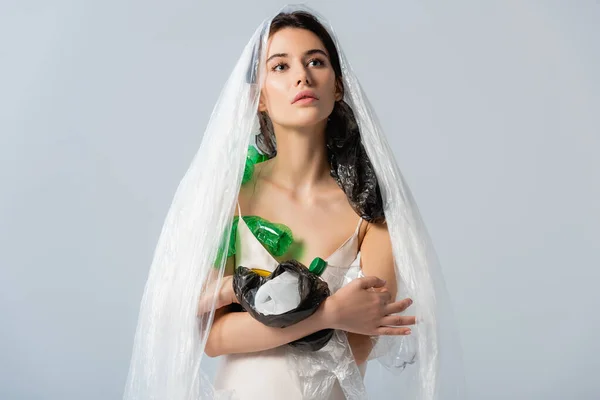 Mujer joven con bolsa de plástico en la cabeza de pie en vestido de seda con botellas vacías aisladas en gris, concepto de ecología - foto de stock