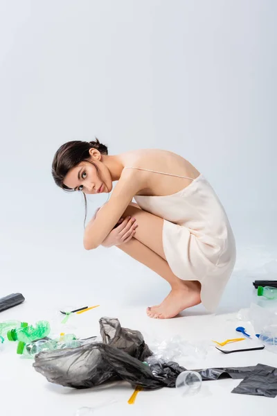 Barfüßige Frau im Seidenkleid, die in die Kamera schaut und neben Plastiktüten und Flaschen auf weißem Grund sitzt, ökologisches Konzept — Stockfoto