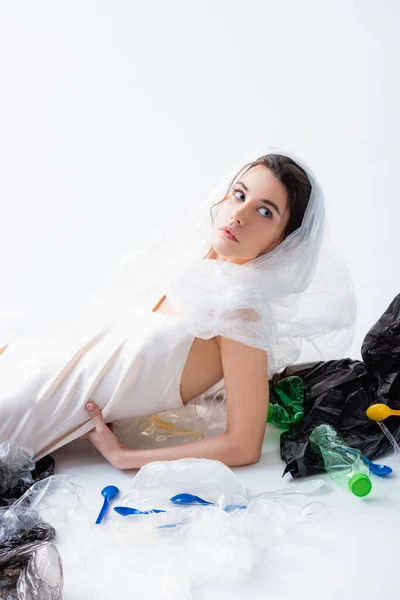 Donna in abito di seta con sacchetto di plastica sulla testa seduta vicino a bottiglie vuote e guardando altrove sul bianco, concetto di ecologia — Foto stock