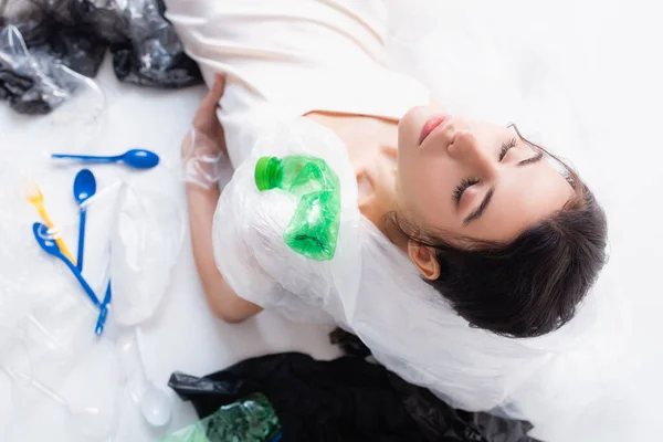 Vista dall'alto della donna con sacchetto di plastica sulla testa e occhi chiusi seduti vicino a bottiglie vuote e cucchiai su bianco, concetto di ecologia — Foto stock