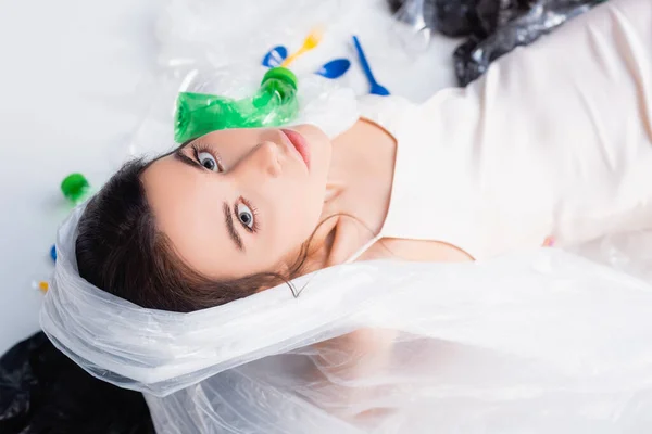 Vista dall'alto della donna con sacchetto di plastica sulla testa seduto vicino a bottiglie vuote e cucchiai su bianco, concetto di ecologia — Foto stock