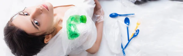 Imagem horizontal da mulher sentada perto de garrafas vazias, sacos de plástico e colheres em branco, conceito de ecologia — Fotografia de Stock