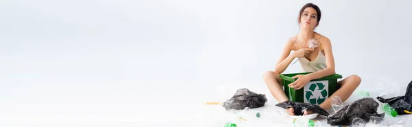 Concepto panorámico de mujer descalza sentada cerca de la papelera con signo de reciclaje mientras sostiene la flor en blanco, concepto de ecología - foto de stock