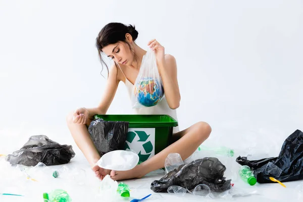 Brunette woman holding plastic bag with globe near rubbish and trash bin with recycle sign on white — Stock Photo
