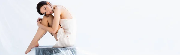 Panoramic crop of barefoot woman in silk dress sitting on stool wrapped in plastic bag on white, ecology concept — Stock Photo