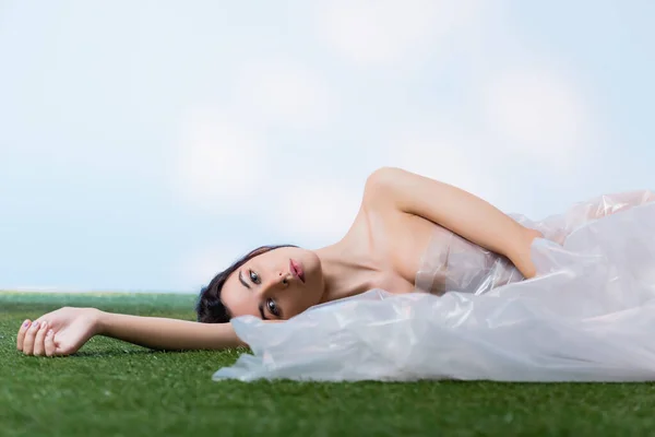 Junge brünette Frau in Polyethylen gehüllt auf grünem Gras liegend und in die Kamera blickend auf blauem, ökologischem Konzept — Stockfoto