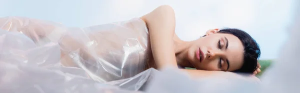 Panoramische Ernte einer brünetten Frau mit geschlossenen Augen, die in Polyethylen gehüllt auf blauem Grund liegt, ökologisches Konzept — Stockfoto