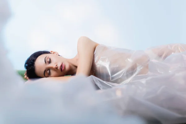Selective focus of brunette woman with closed eyes wrapped in polyethylene lying on blue, ecology concept — Stock Photo
